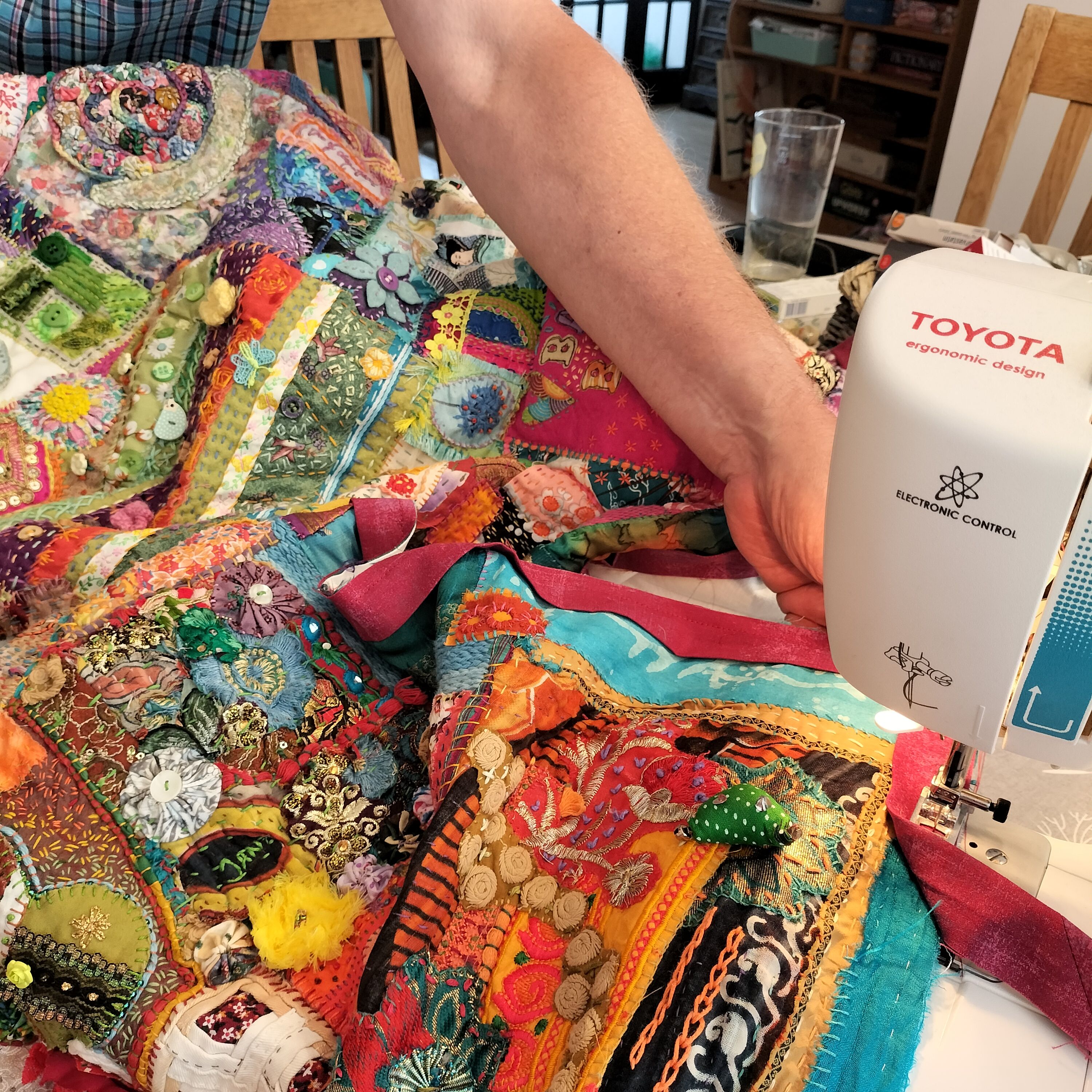 A colourful quilt on a table, with a man's arm holding it in place as it goes through a sewing machine.