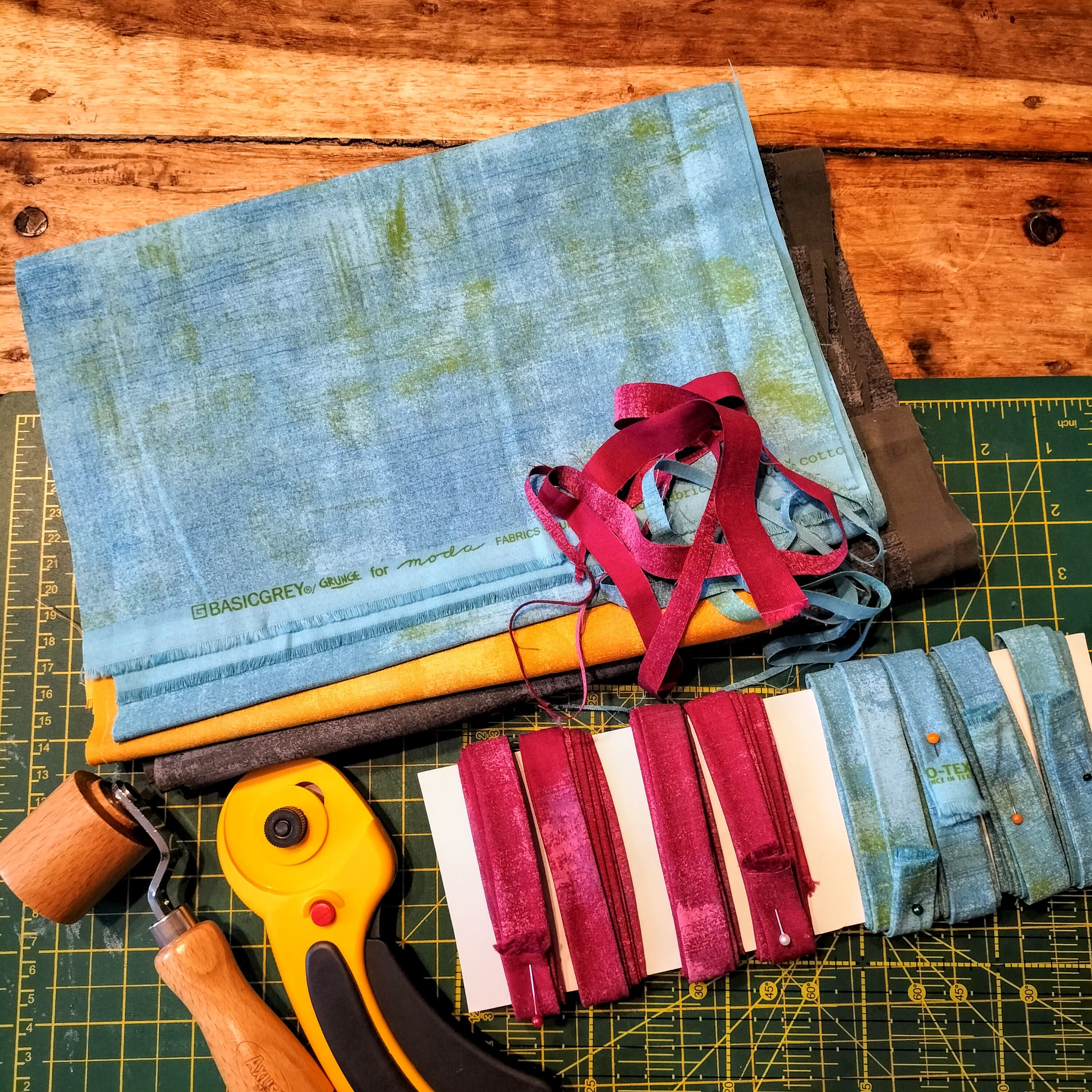 Fabrics, a rotary cutter, seam roller, and strips of binding on a cutting mat.
