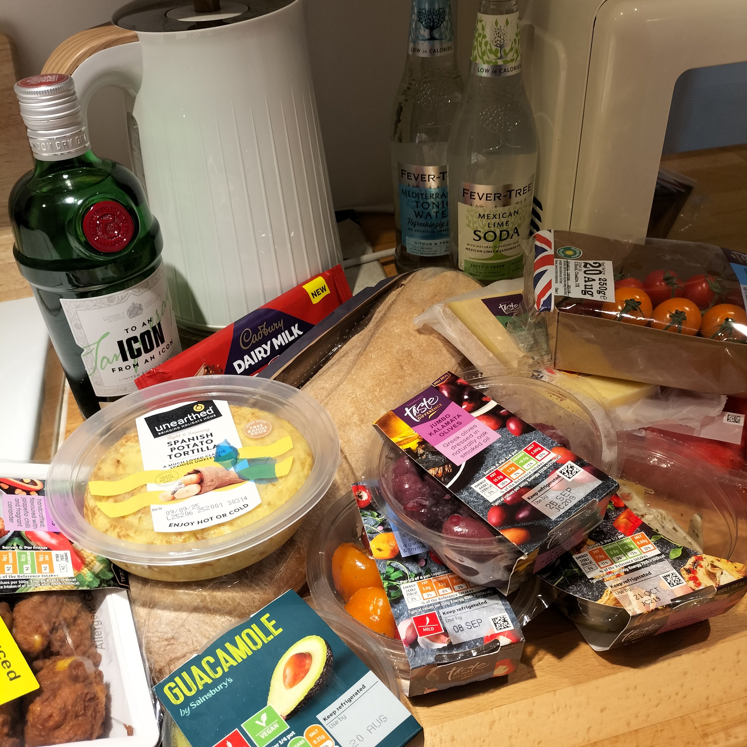 A selection of deli-type foods on a kitchen counter, with a bar of chocolate, some gin and tonics and a kettle in the background